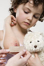 Eine kleines M�dchen bekommt ein Pflaster auf eine Sch�rfwunde am Arm. Sie h�lt einen Teddyb�r fest.