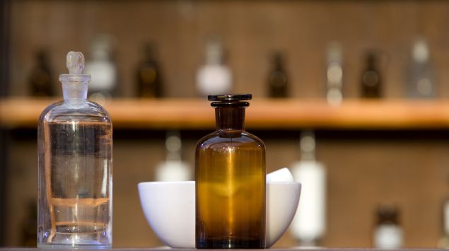 Auf einem Tisch vor einem regal stehen altmodische Apothekerflaschen und ein Mörser.