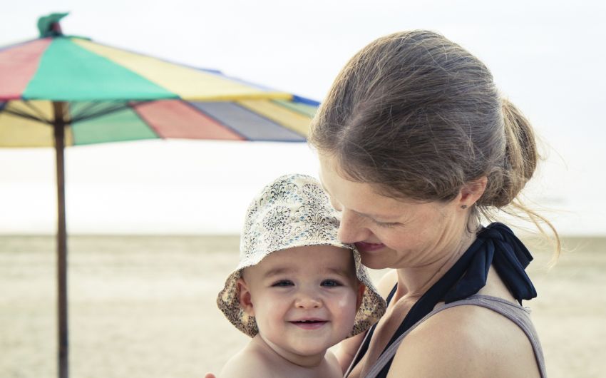 Eine Mutter hält am Strand ihr Baby auf dem Arm, das einen Sonnenhut auf hat.