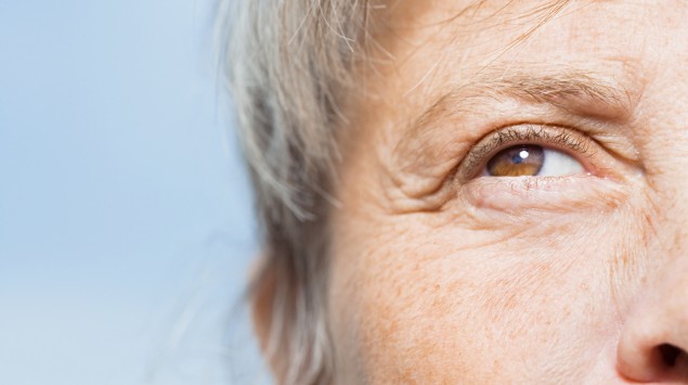 Zu sehen ist ein braunes Auge einer grauhaarigen Person in Nahaufnahme vor blauem Himmel.