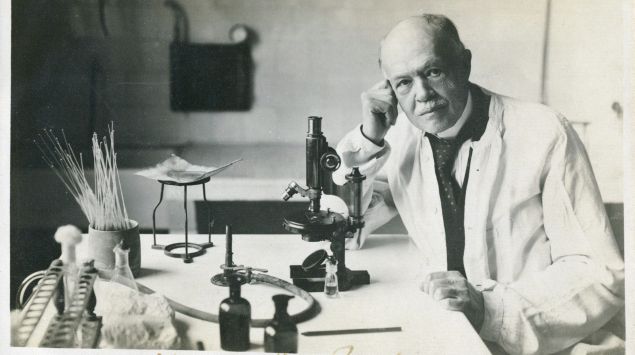 Charles Nicolle sitzt an einem Labortisch, auf dem unter anderem ein Mikroskop steht.