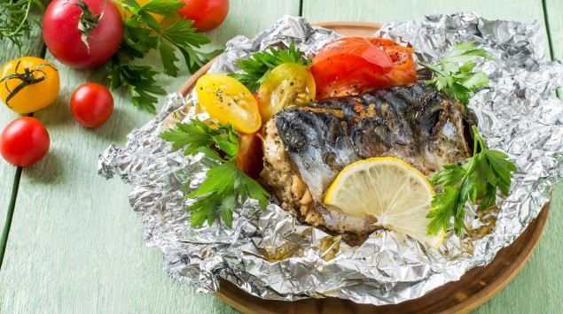 Auf einem Tisch steht ein Teller mit in Alufolie gebackenem Fisch, Tomaten, Zitronenscheiben und Petersilie.