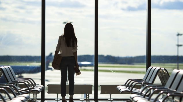 Eine Frau steht im Abflugbereich eines Flughafens und blickt durch das Fenster auf das Flugfeld.