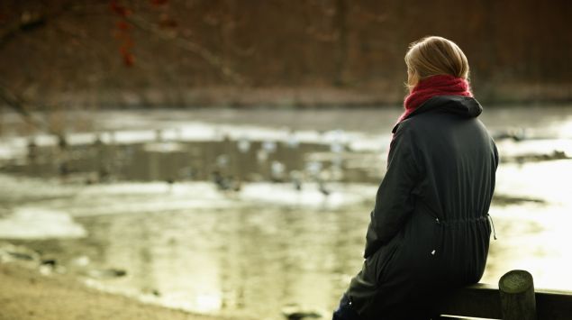Eine winterlich gekleidete Frau sitzt auf der Lehne einer Bank am Ufer eines Sees.