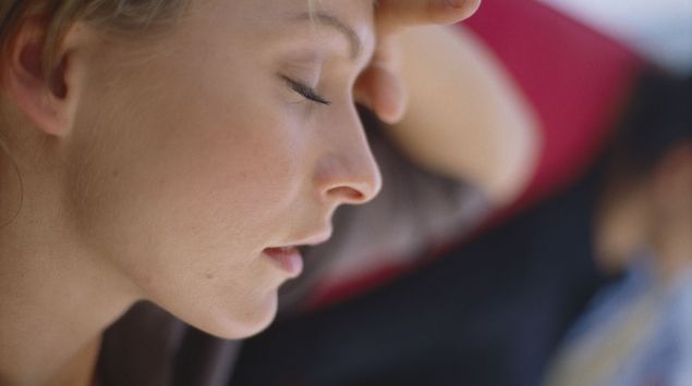 Eine erschöpft wirkende Frau hat die Augen geschlossen und drückt eine Hand gegen die Stirn.