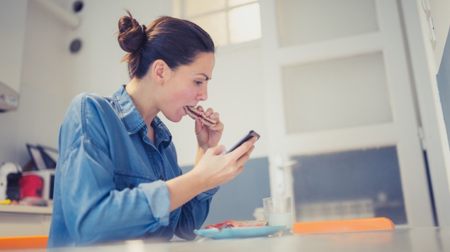 Eine Frau sitzt an einem Tisch und guckt auf ihr Smartphone, während sie ein Brot isst.