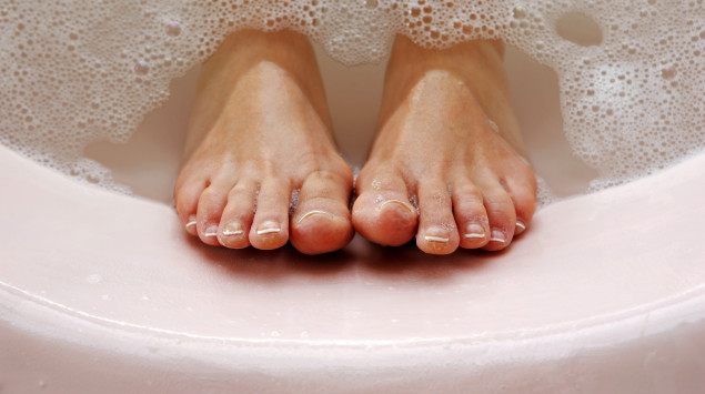 Füße in einer rosa Badewanne mit Schaum im Wasser.