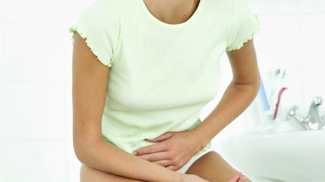 Das Bild zeigt eine junge Frau, die auf dem Bett sitzt und ihren Bauch hält.
