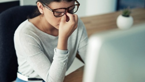 Eine Frau sitzt am PC und greift sich an die Nasenwurzel.