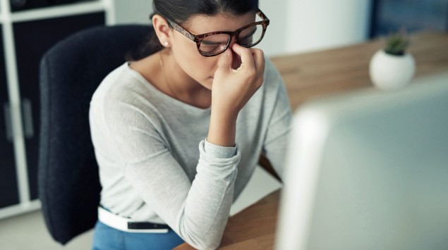 Eine Frau sitzt am PC und greift sich an die Nasenwurzel.