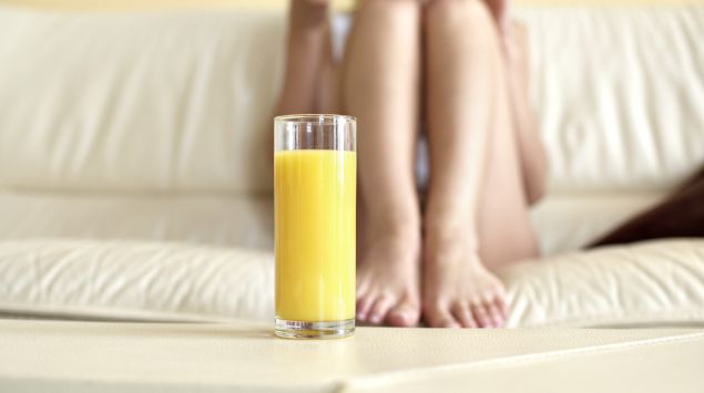 Das Bild zeigt ein Glas mit Orangensaft. Im Hintergrund die Beine einer Frau.