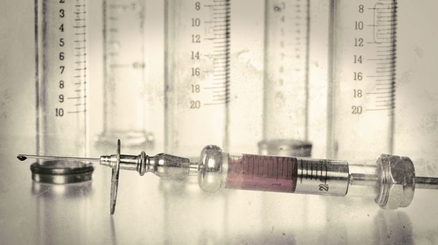 Vor einer Reihe nebeneinanderstehender leerer Glaszylinder liegt eine alte, mit Blut gefüllte Glasspritze.