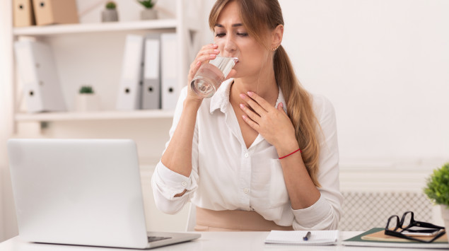 Störender Schleim im Rachen: Eine Frau trinkt ein Glas Wasser und fasst sich an den Hals.