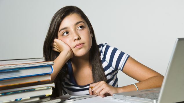 Ein Mädchen sitzt an einem Tisch, auf dem sich ein Laptop, ein Bücherstapel und Schreibblöcke befinden, und blickt in die Luft.