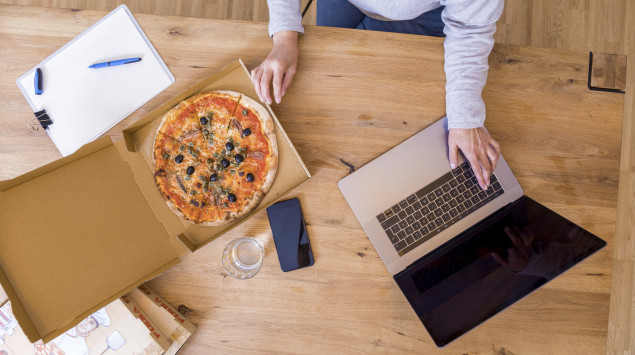 Eine Person sitzt mit Laptop und Pizza am Schreibtisch.