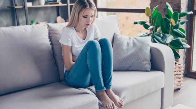 Eine junge Frau mit Regelschmerzen sitzt zusammengekauert auf dem Sofa.