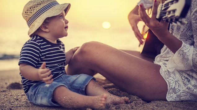 Ein Baby lauscht am Strand seiner Mutter, die Gitarre spielt.