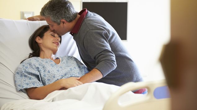 Man sieht einen Mann, der eine Frau im Krankenbett besucht und ihr einen Kuss auf die Stirn gibt.
