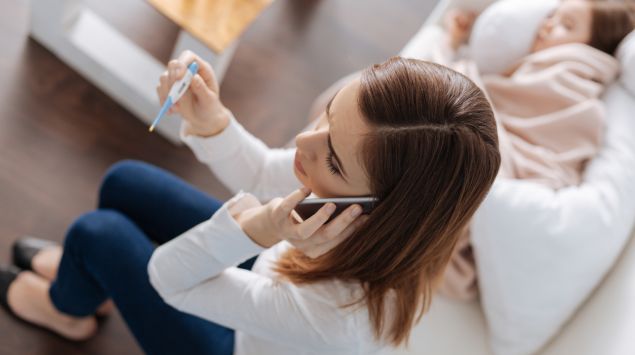 Eine Frau sitzt neben einem kranken Kind auf dem Sofa, hält ein Fieberthermometer in der Hand und telefoniert.
