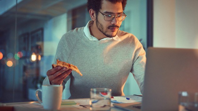 Ein Mann sitzt spätabends am Laptop und hält ein Stück Pizza in der Hand.