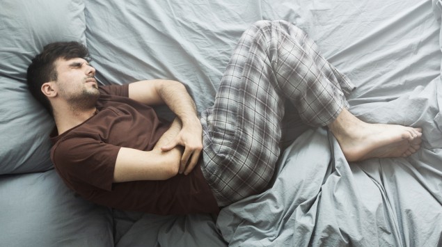 Kolik: Ein Mann liegt mit starken Bauchschmerzen im Bett und hat eine Schonhaltung mit angezogenen Beinen eingenommen.