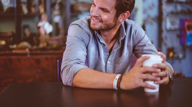 Das Bild zeigt einen Mann, der mit einem Kaffeebecher in der Hand am Tisch sitzt.