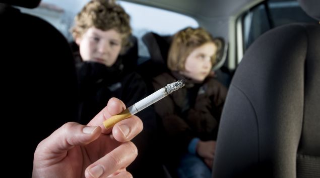 Zwei auf dem Rücksitz eines Autos sitzende Kinder sind dem Zigarettenqualm des rauchenden Beifahrers ausgesetzt.