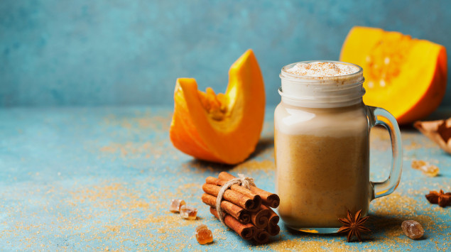 Ein Glas mit Pumpkin Spice Latte, Zimtstangen und Kürbissen im Hintergrund.