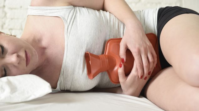 Eine junge Frau mit Schmerzen und Wärmflasche auf dem Bett.