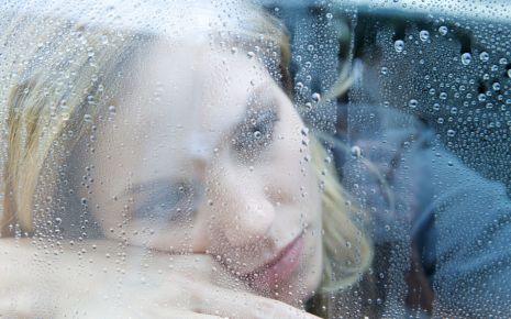 Eine depressiv aussehende Frau schaut durch ein mit Regentropfen bedecktes Fenster.