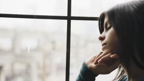 Das Bild zeigt eine junge Frau, die traurig aus dem Fenster guckt.