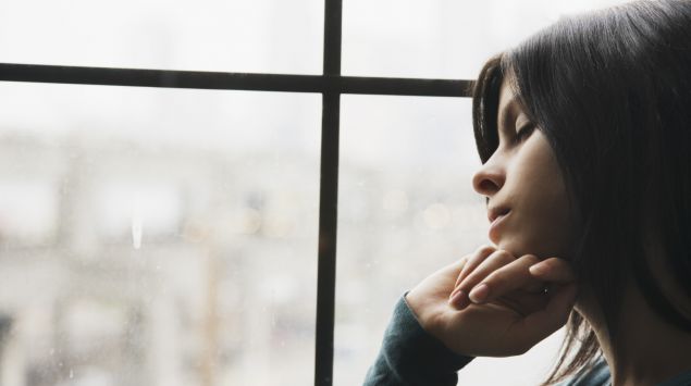 Das Bild zeigt eine junge Frau, die traurig aus dem Fenster guckt.