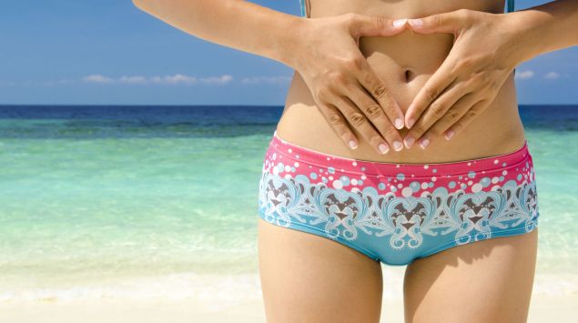 Der Ausschnitt einer Frau im Bikini, die am tropischen Strand steht und die Hände auf den Bauch hält.