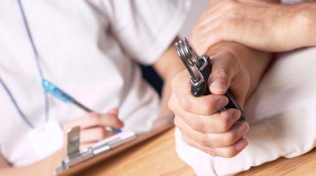 Man sieht eine Hand mit einem Handtrainer und im Hintergrund eine Ärztin mit Klemmbrett und Stift.