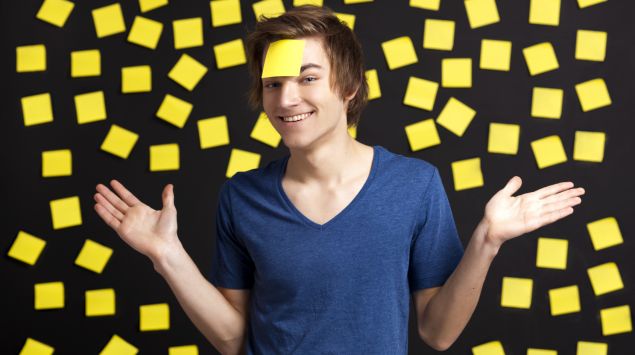 Das Bild zeigt einen jungen Mann vor einer Wand mit Post-its, von denen eines auf seiner Stirn klebt.
