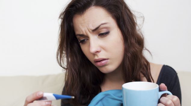 Eine krank aussehende Frau hält eine Tasse und ein Fieberthermometer in der Hand.