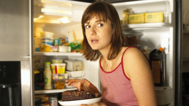 Eine Frau vor dem geöffneten Kühlschrank, sie hält einen Teller mit Schokoladenkuchen in der Hand.