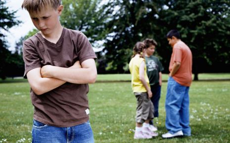 Ein Junge steht einsam und mit verschränkten Armen auf einer Wiese, im Hintergrund eine Gruppe Kinder.