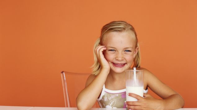 Ein kleines Mädchen sitzt am Tisch und hat ein Glas Milch in der Hand.