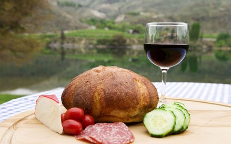 Man sieht einen Laib Brot, Käse, Tomaten, Gurken, Salami und ein Glas Rotwein.