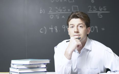 Ein junger Mann sitzt mit Büchern am Tisch, auf der Tafel hinter ihm mathematische Formeln.