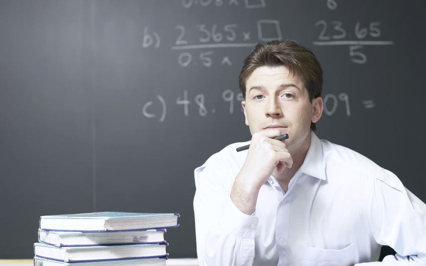 Ein junger Mann sitzt mit Büchern am Tisch, auf der Tafel hinter ihm mathematische Formeln.