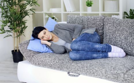 Eine nachdenklich aussehende Frau liegt mit angezogenen Beinen auf einem Sofa.