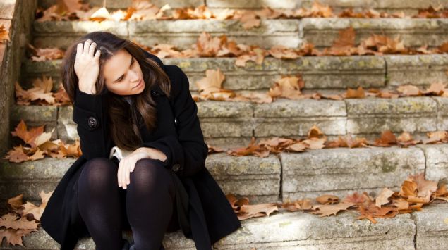 Eine nachdenklich aussehende Frau sitzt auf einer mit herbstlichen Blättern bedeckte Treppe.