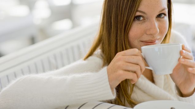 Eine Frau sitzt teetrinkend auf dem Sofa und dreht sich nach hinten zur Rückenlehne.
