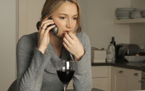 Eine Frau sitzt telefonierend am Küchentisch und hat vor sich ein Glas Wein.