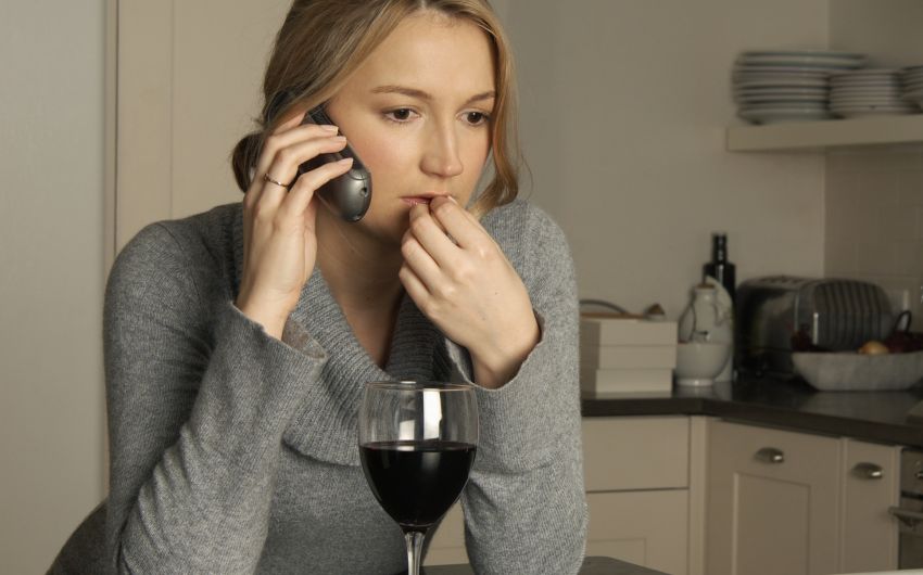 Eine Frau sitzt telefonierend am Küchentisch und hat vor sich ein Glas Wein.
