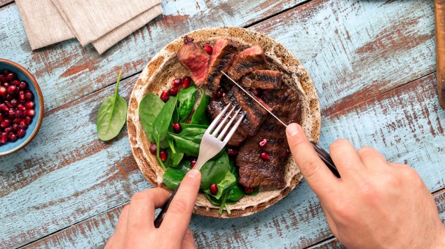 Jemand isst ein gegrilltes Rindersteak mit Salat auf einem blauen Holztisch.