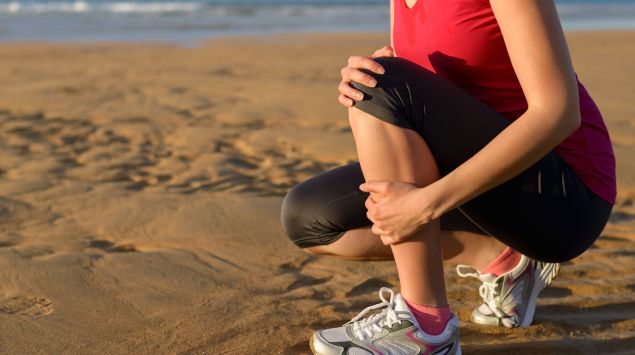 Eine Joggerin kniet am Strand und hält ihr schmerzendes Schienbein.