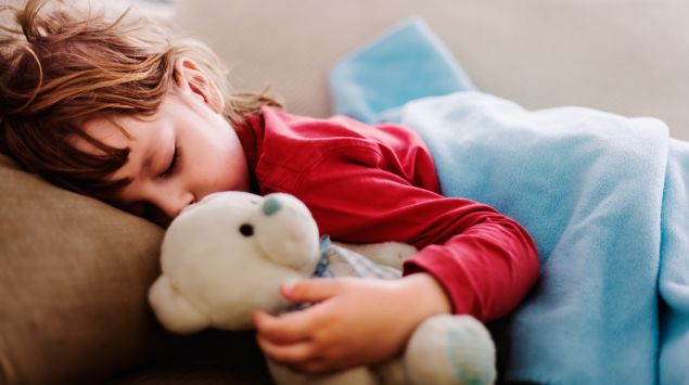 Ein Kind schläft auf einer Couch und hält einen Teddy im Arm.
